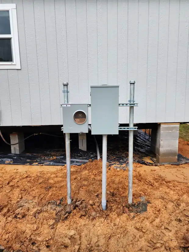 Meter base and disconnect on two galvanized posts beside a raised house, red clay below